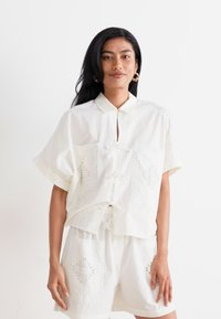 Set di camicia in cotone bianco e pantaloni corti con maniche corte, colletto alla cinese, dettaglio con nodo frontale e ricami sui taschini.