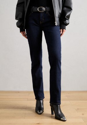 Jeans de mezclilla azul oscuro de corte ajustado, con costuras de contraste. Combinados con botas de tacón negras y una hebilla de cinturón redonda.