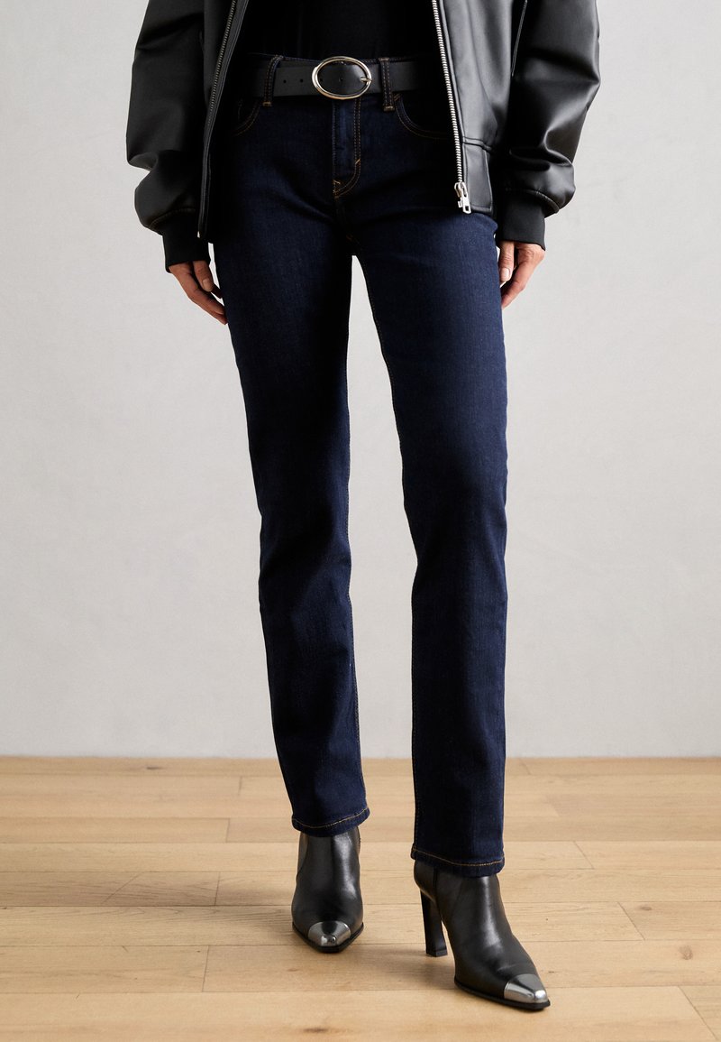 Jeans de mezclilla azul oscuro de corte ajustado, con costuras de contraste. Combinados con botas de tacón negras y una hebilla de cinturón redonda.