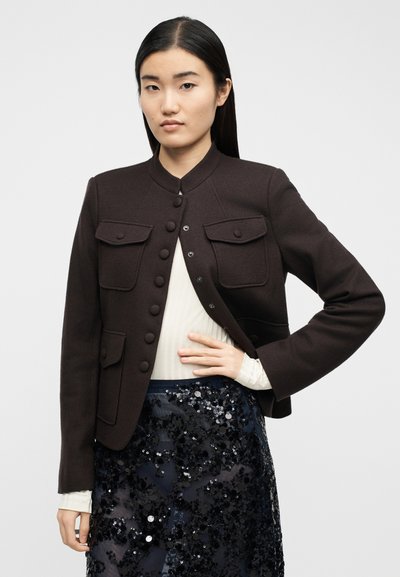 Γυναίκα με μακριά σκούρα μαλλιά, φοράει ένα σκούρο καφέ σακάκι με κουμπιά πάνω από ένα λευκό ριπτό τοπ και μια μαύρη φούστα με παγιέτες, στέκεται με το ένα χέρι στη μέση.