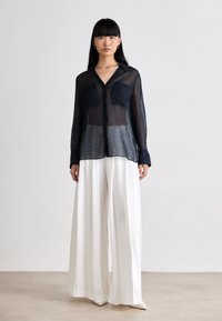 Camisa azul-marinho transparente com dois bolsos frontais, combinada com calças brancas de pernas largas, apresenta mangas longas e um colarinho clássico.