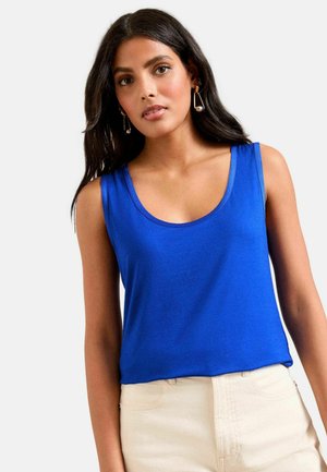 Frau mit langem dunklem Haar, die baumelnde Ohrringe, ein ärmelloses, königsblaues Oberteil und hellbeige, hoch taillierte Hosen trägt.