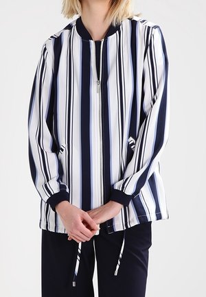 Person wearing a long-sleeve, vertical striped zip-up jacket in navy, white, and light blue, with hands clasped in front.