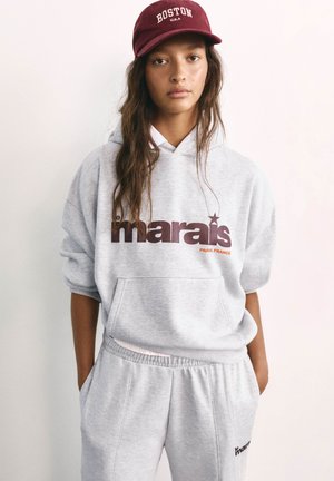 Ung person iført en grå "Le Marais Paris France" hættetrøje og matchende joggingbukser, med en bordeaux Boston kasket, står med hænderne i lommerne.