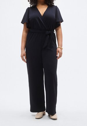 Femme portant une combinaison noire portefeuille à manches courtes avec une ceinture à la taille, associée à des chaussures à talons beige et un bracelet en or.
