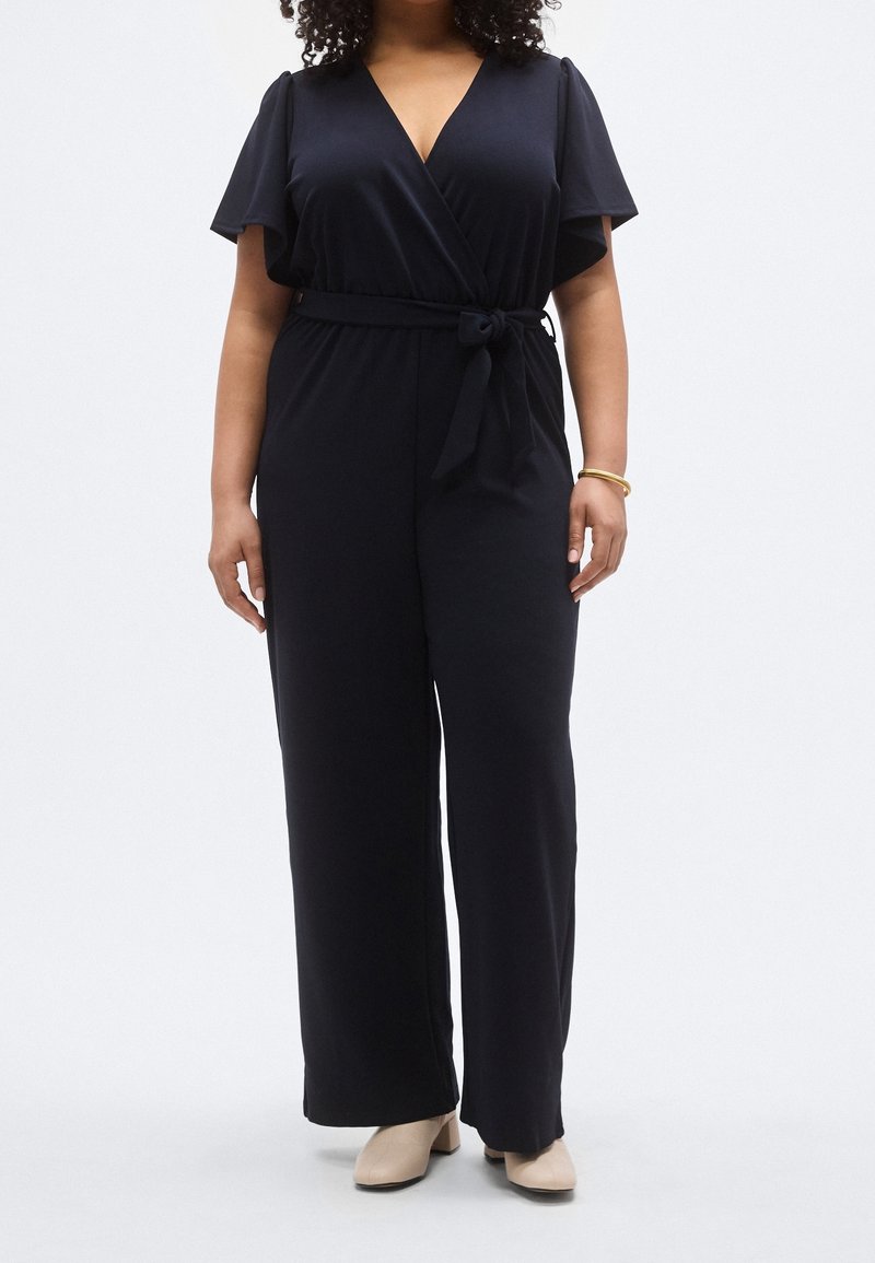 Femme portant une combinaison noire portefeuille à manches courtes avec une ceinture à la taille, associée à des chaussures à talons beige et un bracelet en or.
