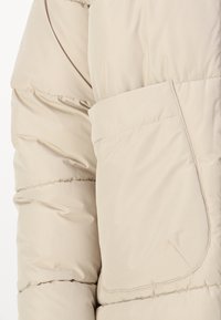 Doudoune beige avec une surface texturée, des coutures horizontales et une poche à rabat sur le bras gauche.