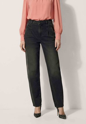 Vrouw die zwarte high-waist jeans draagt, een roze blouse met lange mouwen en patronen op haar hoge hakken, staand tegen een lichte achtergrond.