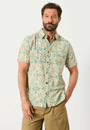 Mann mit Bart, der ein kurzärmliges Hemd mit grün-beigem geometrischem Muster und beige Cargoshorts trägt, steht vor weißem Hintergrund.
