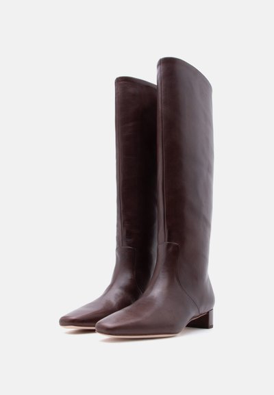 Ψηλές καφέ σκούρες δερμάτινες μπότες με χαμηλά τακούνια και στρογγυλεμένες τετράγωνες μύτες, όρθιες πάνω σε απλό λευκό φόντο.