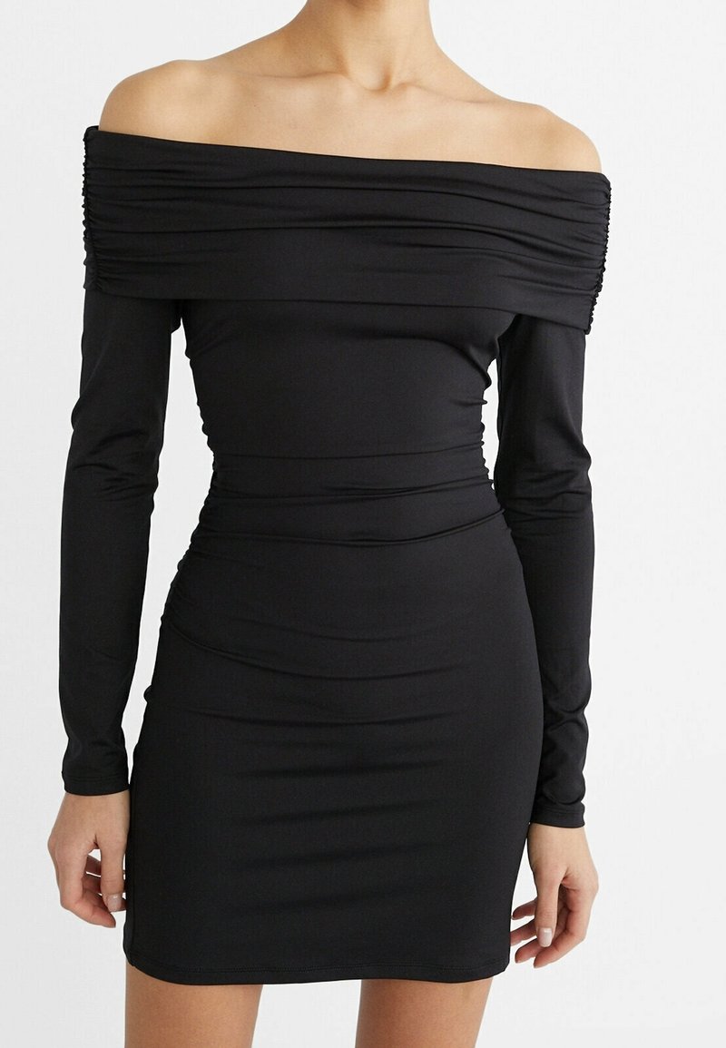 Femme portant une robe noire ajustée épaules dénudées, manches longues, encolure froncée et un ourlet court sur fond blanc.