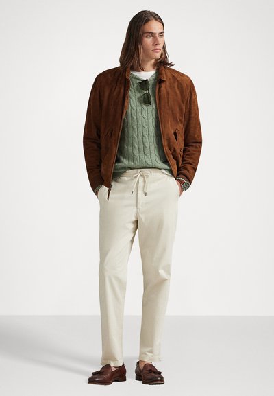 Casaco bomber de camurça castanho por cima de um suéter verde de malha trançada, calças cremes com cordão e sapatos de pele castanha com um detalhe em costura.