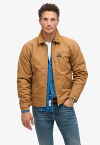 Bruine canvas jas met een ritssluiting aan de voorkant, zijzakken en een borstzak. Draagt over een blauwe spijkerblouse, gecombineerd met blauwe spijkerbroek.