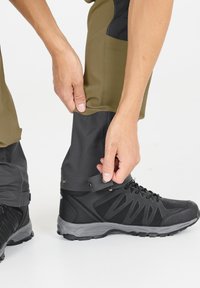 Svarta vandringskängor med grå accenter i kombination med olivgröna och svarta byxor. Byxorna har en tryckknappslåsning vid fållen för justering.