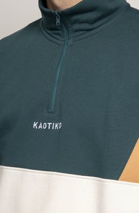 Pullover a mezze zip in teal e crema con accento arancione. Caratterizzato da un tessuto morbido, colletto alto e logo ricamato "KAOTIKO" sul petto.