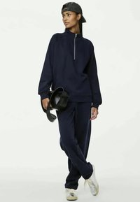 Sweatshirt bleu marine zippé avec des poignets côtelés, associé à un pantalon assorti. Le mannequin tient une casquette noire, portant des baskets argentées. Tissu lisse, coupe décontractée.