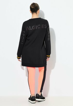 Zwarte lange mouwen tuniek met tekstdetails en oranje accenten, gecombineerd met roze en zwarte kleurgeblocte leggings en zwarte sneakers.