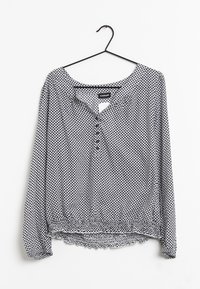 Blusa a maniche lunghe con motivo bianco e nero, orlo elastico e scollatura con bottoni, appesa a una gruccia nera su sfondo bianco.