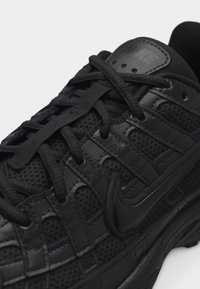 Κοντινό πλάνο ενός μαύρου αθλητικού παπουτσιού Nike που διαθέτει πάνελς από δίχτυ, επικαλύψεις από δέρμα, κορδόνια και το λογότυπο Nike στη πλευρά.