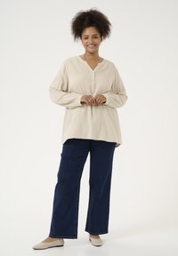 Beige Langarmoberteil mit halber Knopfleiste, kombiniert mit weit geschnittenen dunkelblauen Jeans und hellen Ballerinas, vor einem einfarbigen Hintergrund.