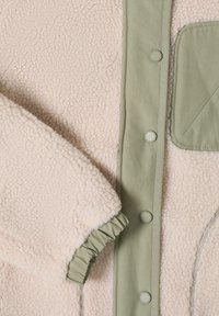 Zbliżenie na beżową kurtkę z polaru typu sherpa z oliwkowozielonym panelem z zatrzaskami, elastycznym mankietem i pasującą kieszenią na piersi.