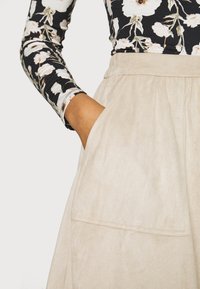 Falda de ante beige con bolsillos laterales y cintura fruncida, combinada con una blusa negra de manga larga con flores. Primer plano de una mano en el bolsillo.