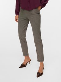 Sieviete, tērpusies pelēkās pielāgotās biksēs un smailos brūnos augstpapēžu zābakos ar metāla caurumu detaļām, stāv ar vienu roku kabatā.