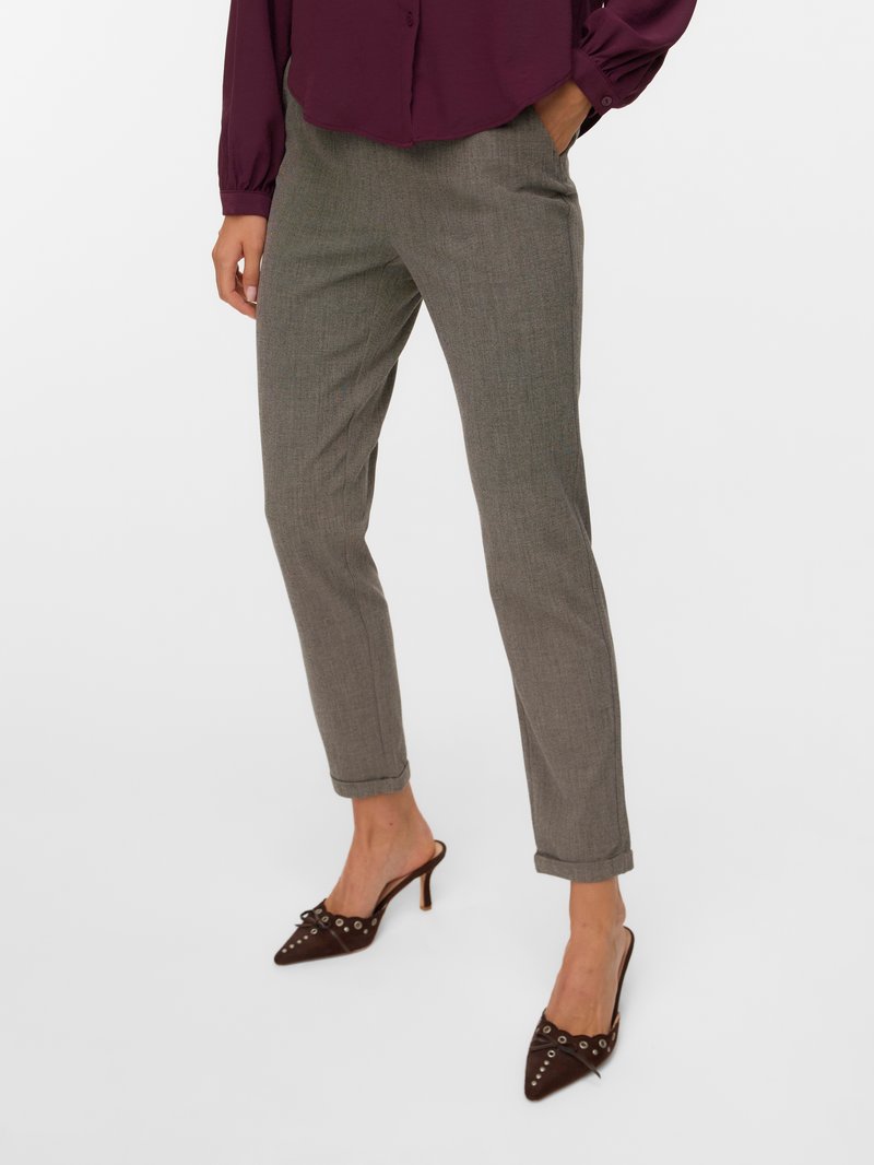 Sieviete, tērpusies pelēkās pielāgotās biksēs un smailos brūnos augstpapēžu zābakos ar metāla caurumu detaļām, stāv ar vienu roku kabatā.