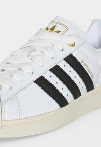Baskets en cuir blanc avec bandes noires, lacets blancs, semelle beige, et texte ainsi que logo dorés "SUPERSTAR" sur la languette.