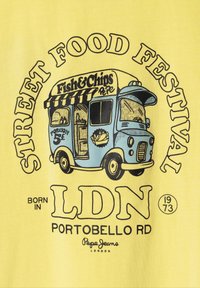 Geltona marškinėliai su mėlynu maisto furgono piešiniu, pažymėtu "Žuvies ir traškučių". Tekste yra "GATVĖS MAISTO FESTIVALIS", "LDN", "PORTOBELLO RD" ir "1973".