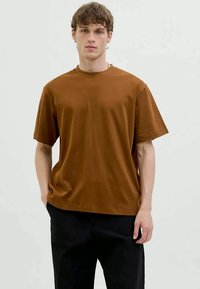 Jovem vestido com uma t-shirt simples de mangas curtas castanha e calças pretas, em pé com uma mão no bolso contra um fundo claro.