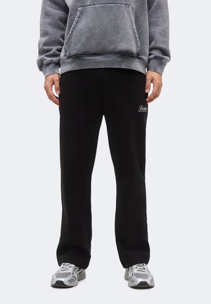 Person iført sorte træningsbukser med et lille hvidt logo, grå hættetrøje og sølvgrå sportssko, stående med afslappede arme langs siden.