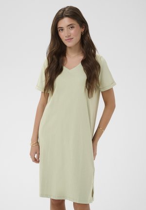 Mujer con cabello largo y castaño, vestida con un vestido de manga corta color verde claro con escote en V y joyería dorada, de pie con las manos relajadas.