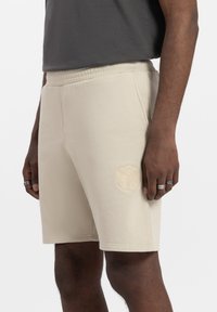 Beige katoenen shorts met een elastische tailleband, zijzakken en een tonale logo-geborduurde opdruk op het linkerpoot. Glad textuur, knielengte ontwerp.
