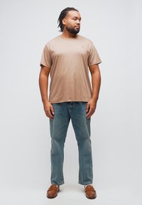 Άνδρας με πλεγμένα μαλλιά, φορώντας καφέ μπλουζάκι, μπλε τζιν και καφέ μοκασίνια, στέκεται και κοιτάζει προς το πλάι μπροστά σε απλό φόντο.