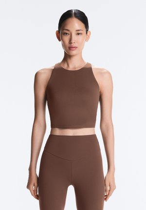 Frau mit dunklem, zurückgebundenem Haar trägt ein ärmelloses braunes Crop-Top und passende hoch taillierte Leggings, stehend vor einem schlichten Hintergrund.