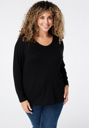 Lachende vrouw met krullend haar, gekleed in een zwart shirt met lange mouwen en blauwe spijkerbroek, staand tegen een effen witte achtergrond.