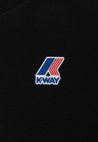 Der schwarze Stoff zeigt ein K-Way-Logo, das aus dem Buchstaben "K" in Blau und Rot mit diagonalen Streifen und abgerundeten Kanten besteht, vor dem Hintergrund der Textur.