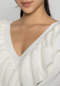 Maglione bianco morbido con una texture a costine, dotato di un profondo scollo a V e di un design arricciato lungo le spalle e il collo.