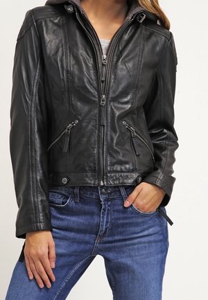 Kvinde iført en sort lynlåset læderjakke med lynlåslommer og blå jeans, hånd hvilende i lommen.