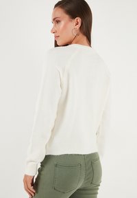 Suéter blanco de punto con cuello redondo, puños y dobladillo acanalados. Lleva puesto un pantalón verde oliva, mostrando un diseño simple y texturizado.