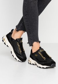 Svarta syntetiska sneakers i mocka med guldkedjesmycken, tjock vit sula och sportig design. Bärs med fransade svarta jeans.