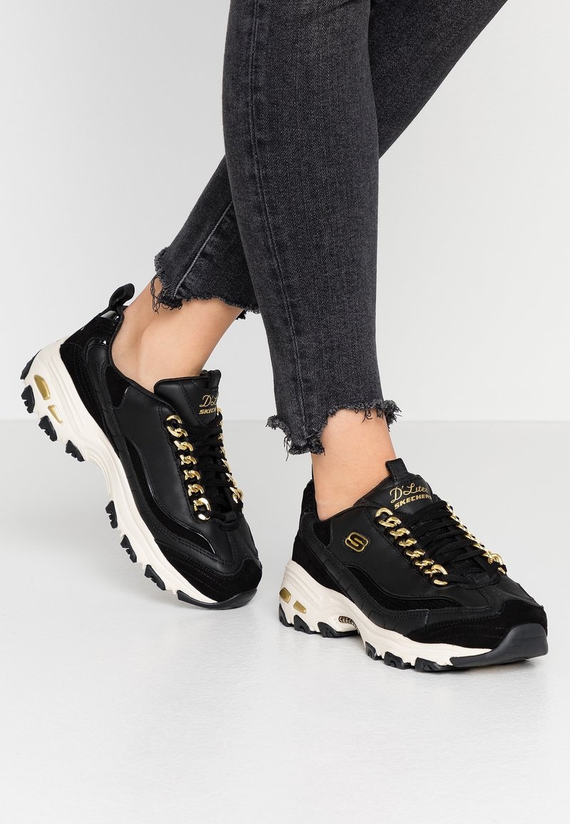 Svarta syntetiska sneakers i mocka med guldkedjesmycken, tjock vit sula och sportig design. Bärs med fransade svarta jeans.