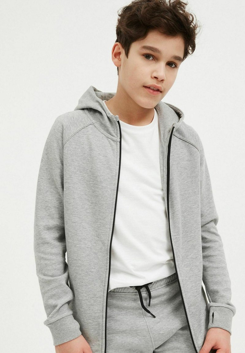 Grauer Reißverschluss-Hoodie mit gefütterter Kapuze, ausgestattet mit schwarzen Akzenten und einer glatten Textur, getragen über einem weißen T-Shirt. Lockere Passform mit langen Ärmeln.
