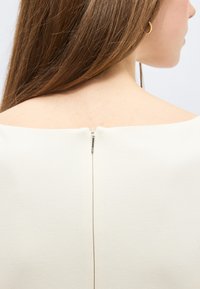 Εγγύτατη λήψη της πλάτης μιας γυναίκας που δείχνει μακριά καστανά μαλλιά, ένα χρυσό κρίκο σκουλαρικιού και τη λεπτομέρεια του φερμουάρ σε ένα λευκό τοπ με ώμο έξω.