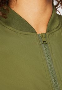 Veste bomber vert olive avec un col côtelé, fermeture éclair à l'avant et texture de tissu lisse. Les détails comprennent une fermeture éclair assortie et des coutures.