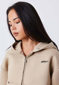 Sudadera con capucha y cremallera beige, con una cremallera plateada y detalle de logo en el pecho, hecha de un tejido suave y liso.
