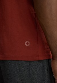 Close-up van de torso van een persoon die een rood shirt draagt met een klein kruislogo nabij de zoom en gestructureerde donkergrijze broek.