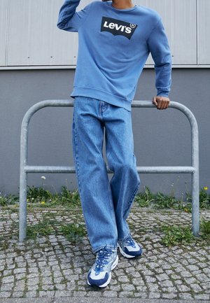 Osoba oparta o metalową poręcz, ubrana w niebieską bluzę Levi's, luźne niebieskie dżinsy i niebiesko-białe trampki na kamiennej nawierzchni z trawą.
