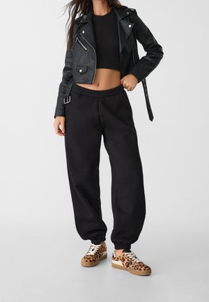 Black cropped leather jacket over a black top, paired with loose black sweatpants and tan sneakers featuring a leopard print design.